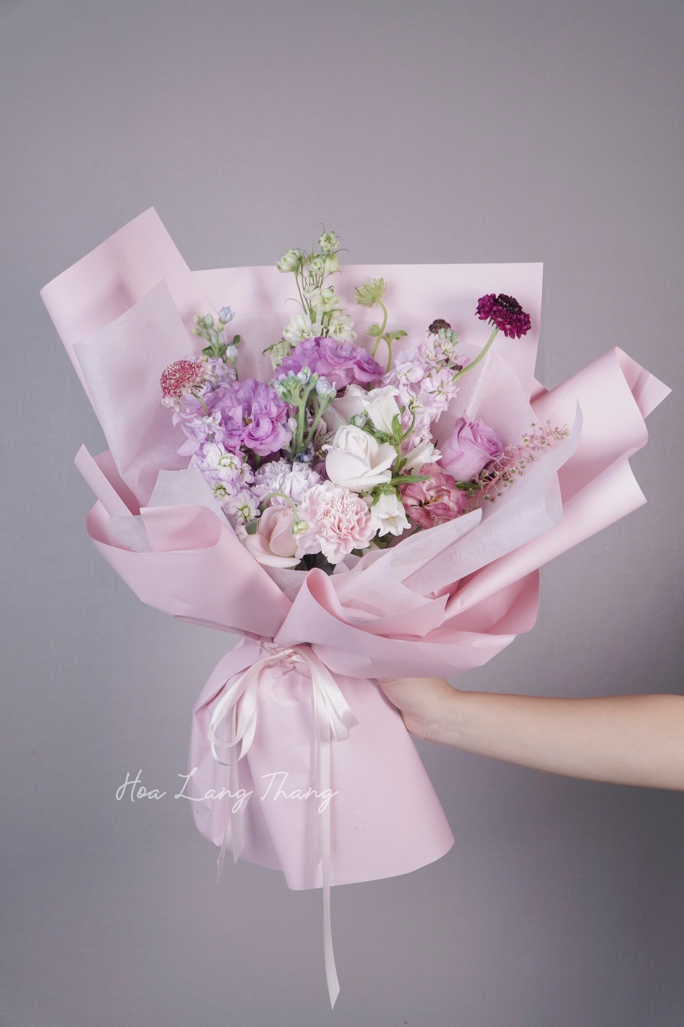Bó hoa sắc màu pastel gồm hoa hồng, lan, và cát tường trong giấy gói hồng ngọt ngào