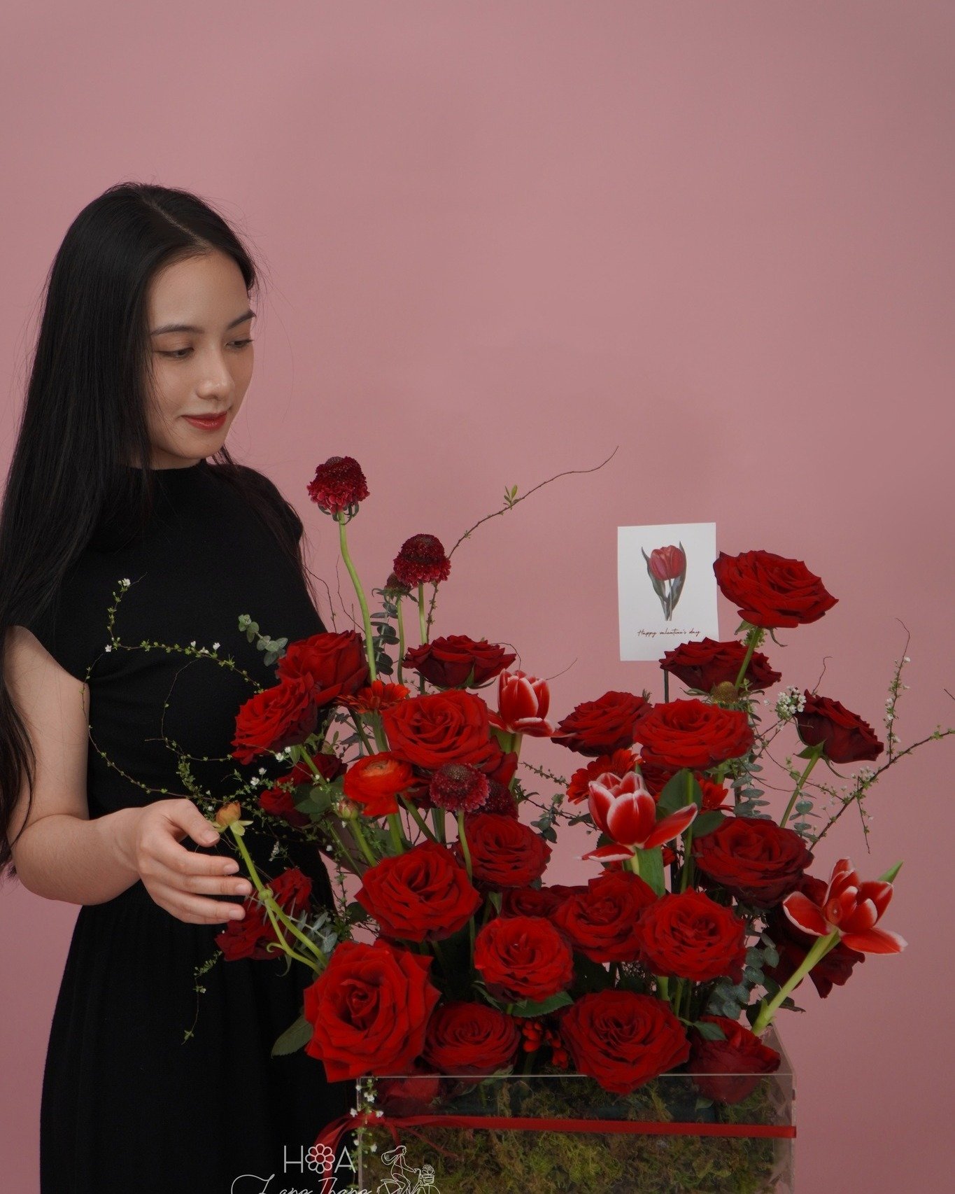 Cô gái đứng gần giỏ hoa hồng đỏ, hoa tulip và các loại hoa khác trên nền hồng
